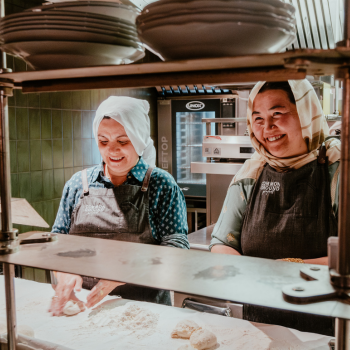 Two women in aprons and headscarves smiling while preparing dough together in a commercial kitchen.